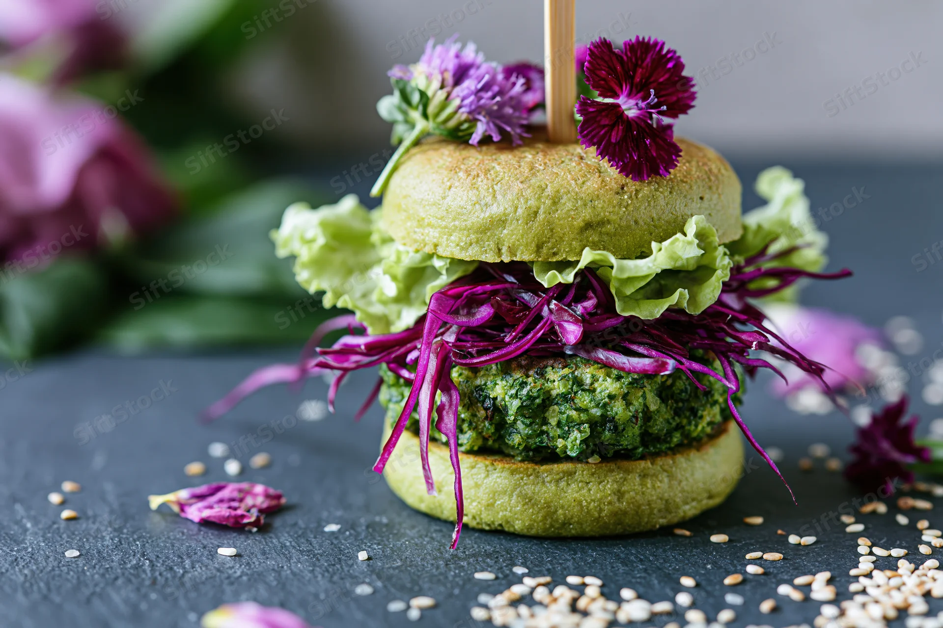 Vegan avocado bun burger with green herb patty and purple cabbage garnished with edible flowers on dark surface