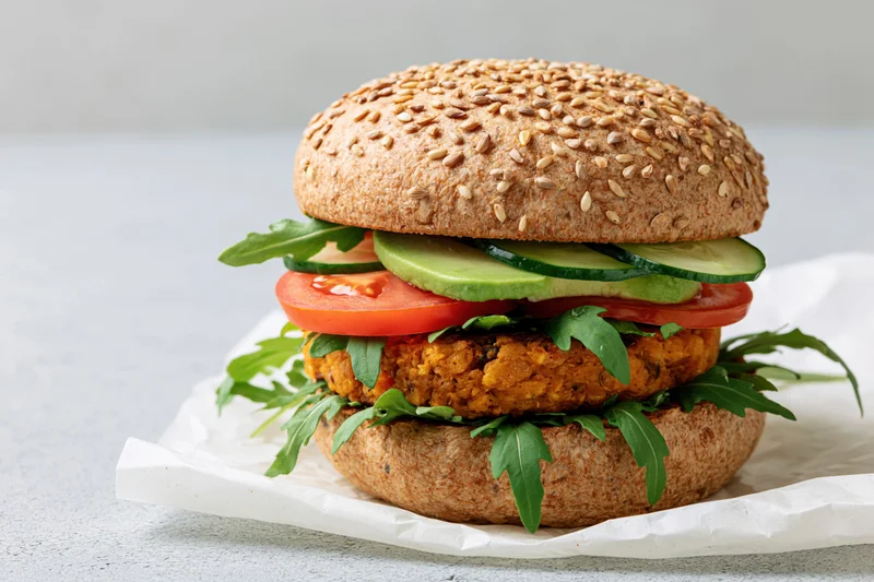 Vegan burger with chickpea patty and fresh vegetables stacked on sesame seed bun on white paper