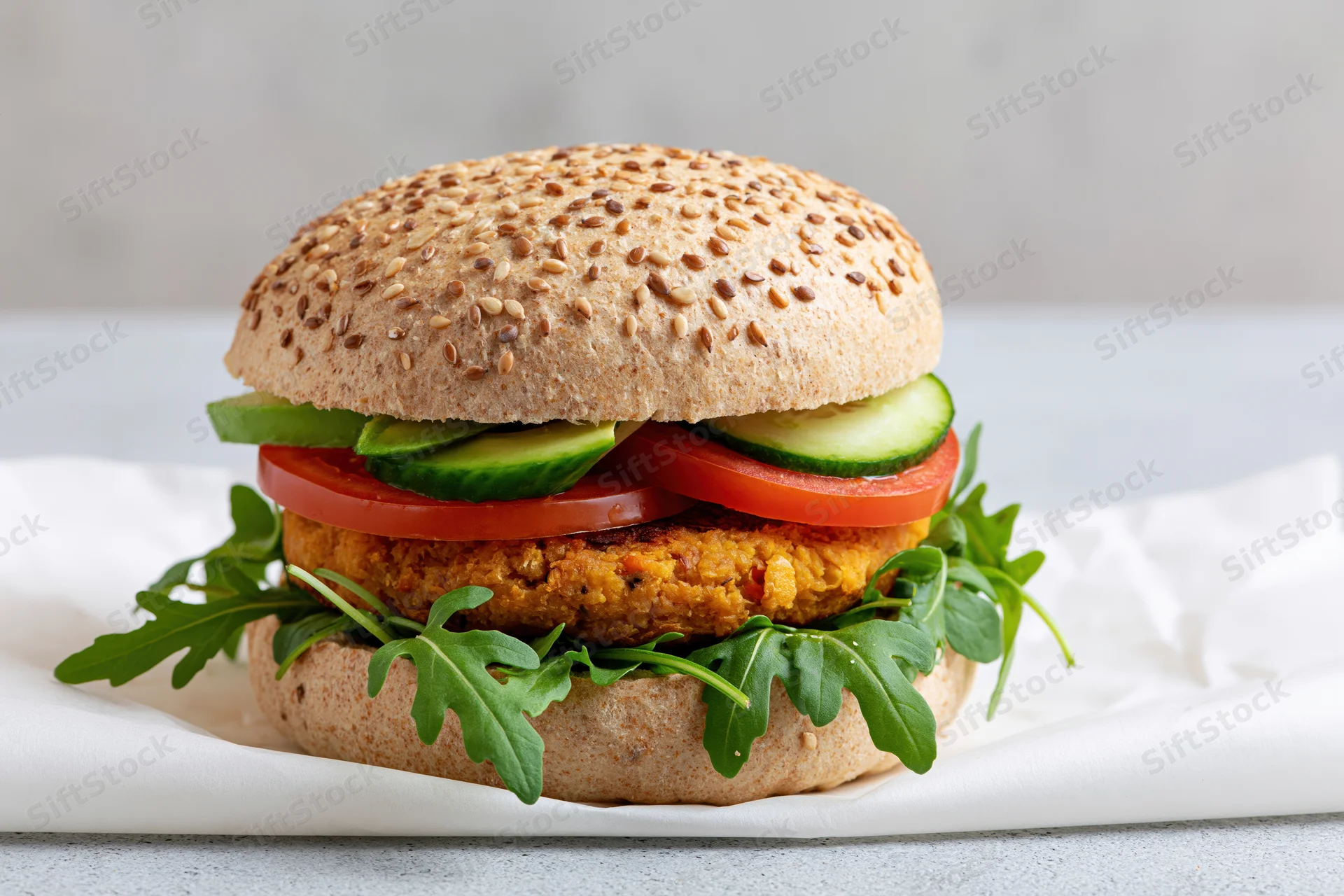 Vegan burger with chickpea patty and fresh vegetables on a seeded bun, resting on a white cloth over a gray surface
