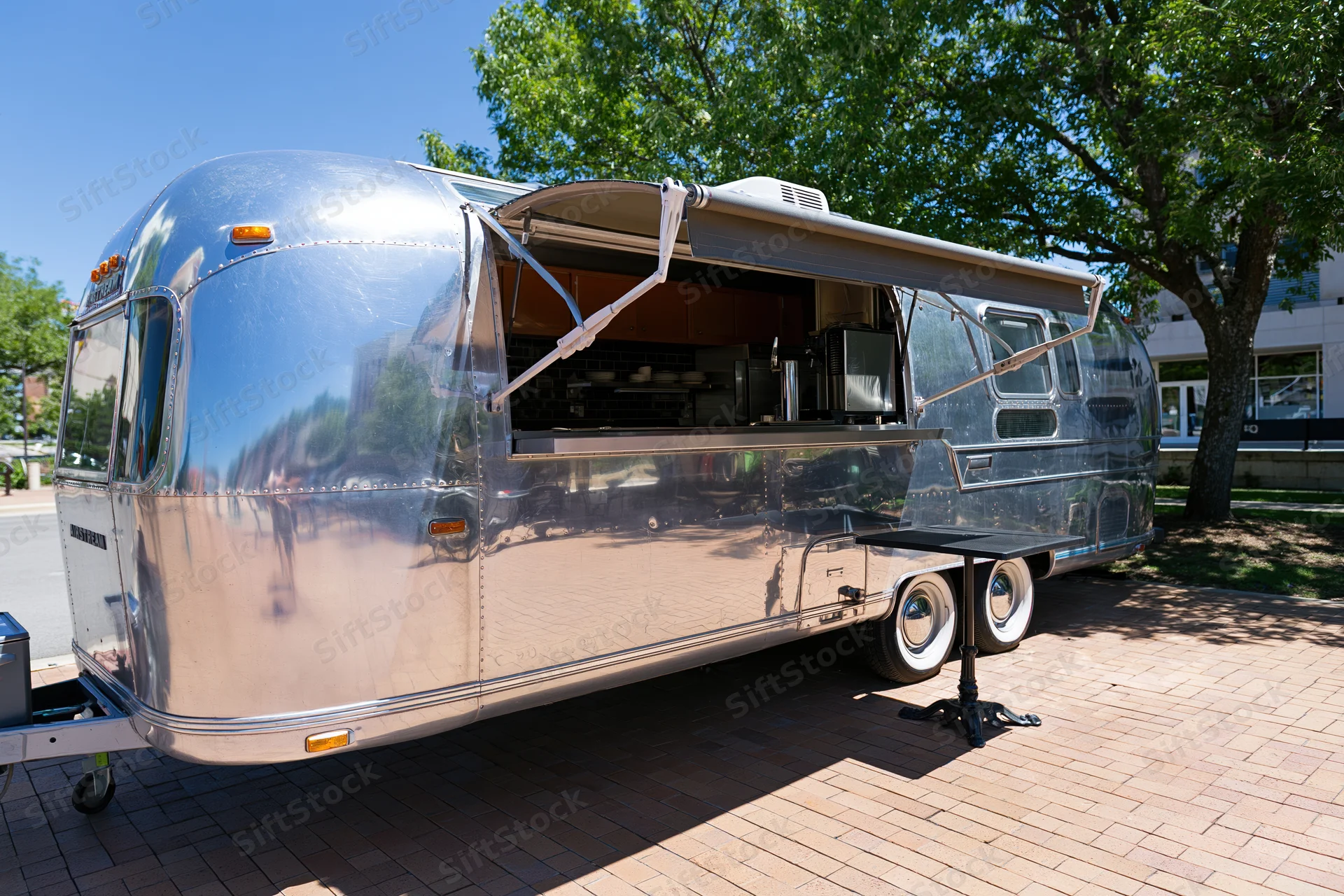 Vintage polished aluminum airstream trailer with serving awning open on brick pavement under tree shade