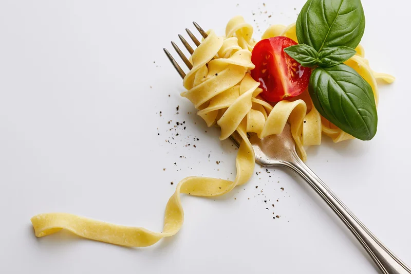 Vintage silver fork holding twisted tagliatelle pasta with basil and tomato on white surface
