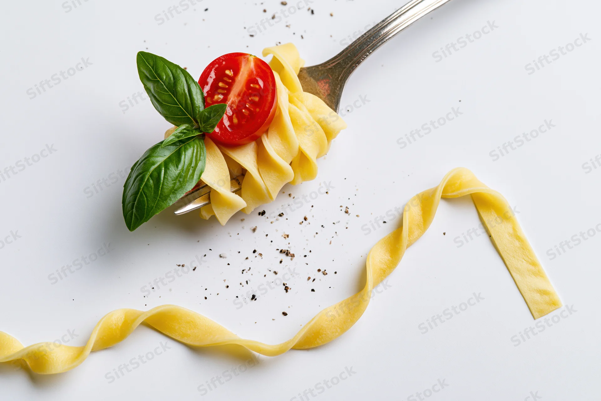 Vintage silver fork holding twisted tagliatelle pasta with cherry tomato and basil leaf on white surface