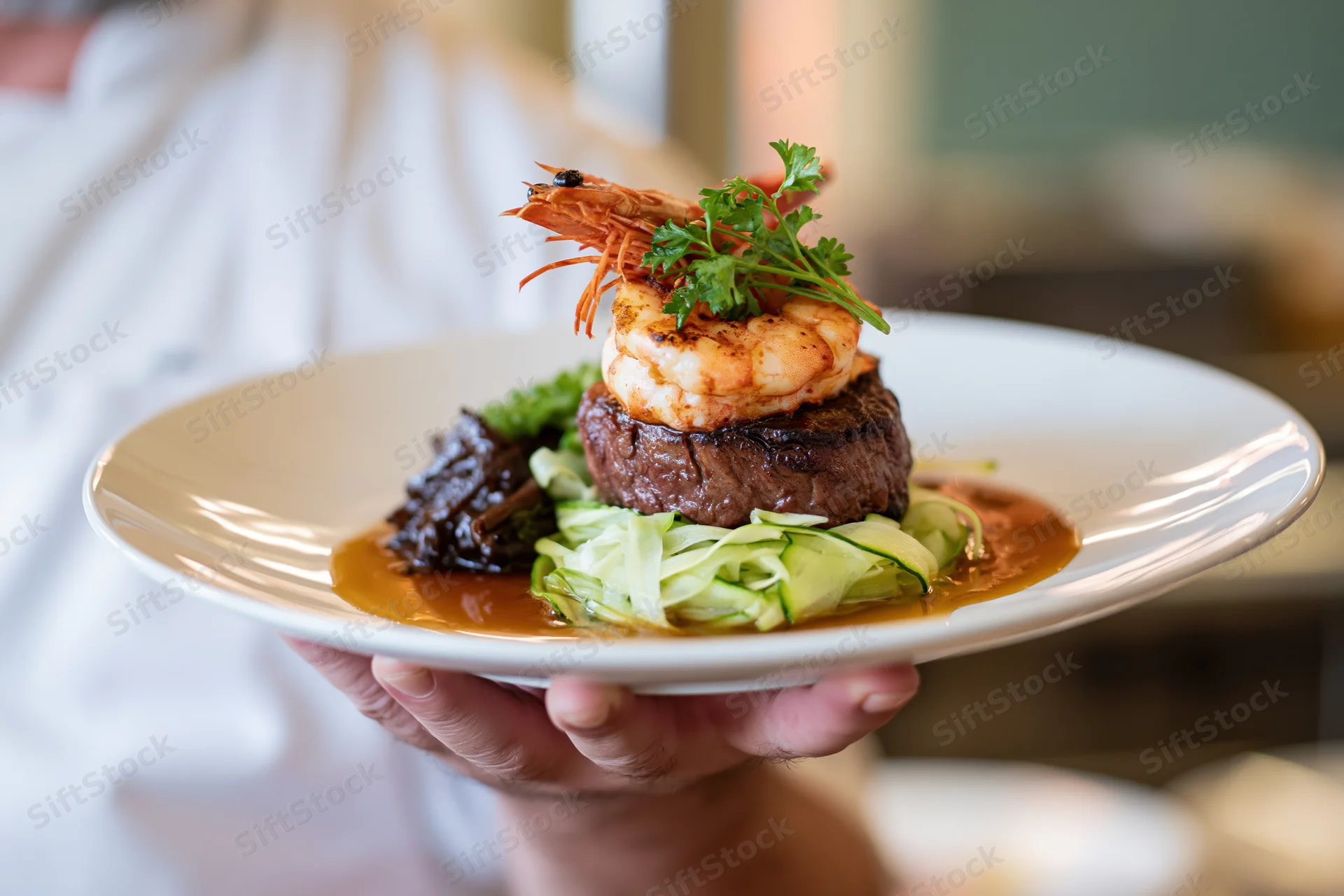 Waiter holding a plated steak topped with shrimp and garnished with parsley and zucchini in a restaurant setting