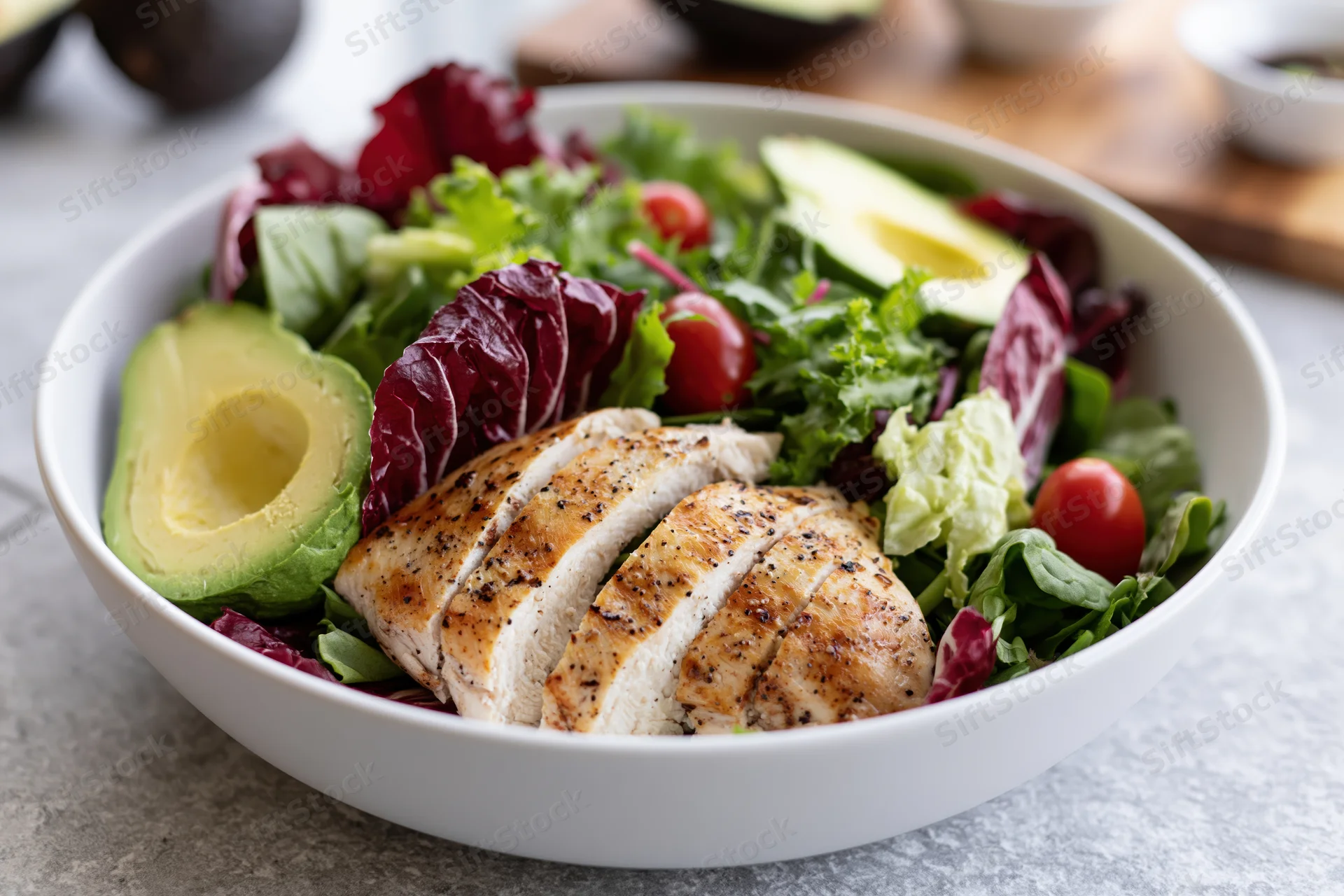 Grilled chicken breast and avocado salad in a white bowl with mixed greens and cherry tomatoes