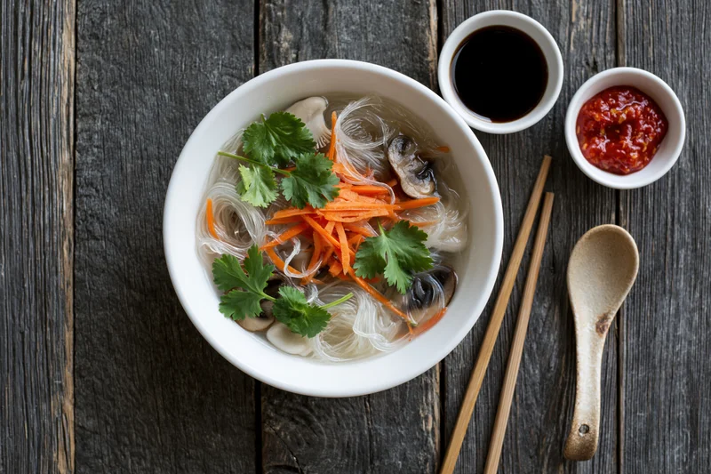 White ceramic bowl of asian noodle soup with glass noodles, carrots, cilantro, mushrooms, chopsticks and spoon on dark wooden table