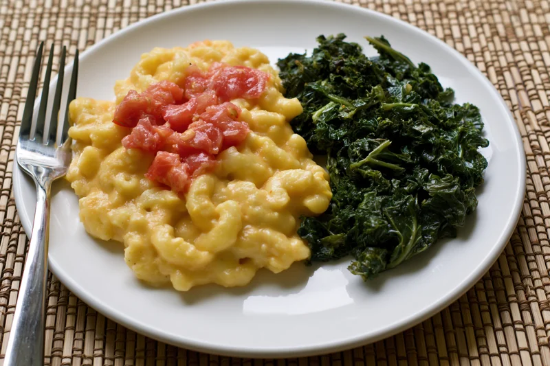 Creamy macaroni and cheese with diced tomato and sautéed kale on a white plate with fork on woven placemat