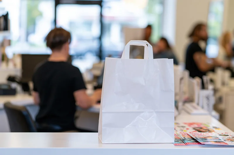 White plastic takeout bag on a white counter in a cafe with blurred staff and customers in the background