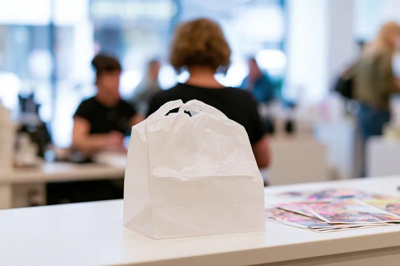 White plastic takeout bag on a white counter in a restaurant interior with blurred staff in background