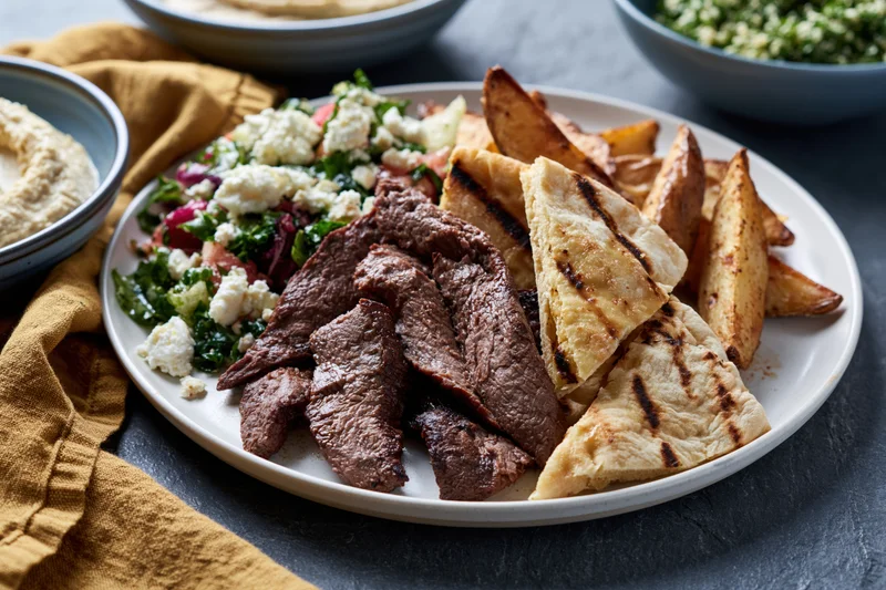 Plate of sliced grilled beef with grilled pita wedges and salad on dark surface