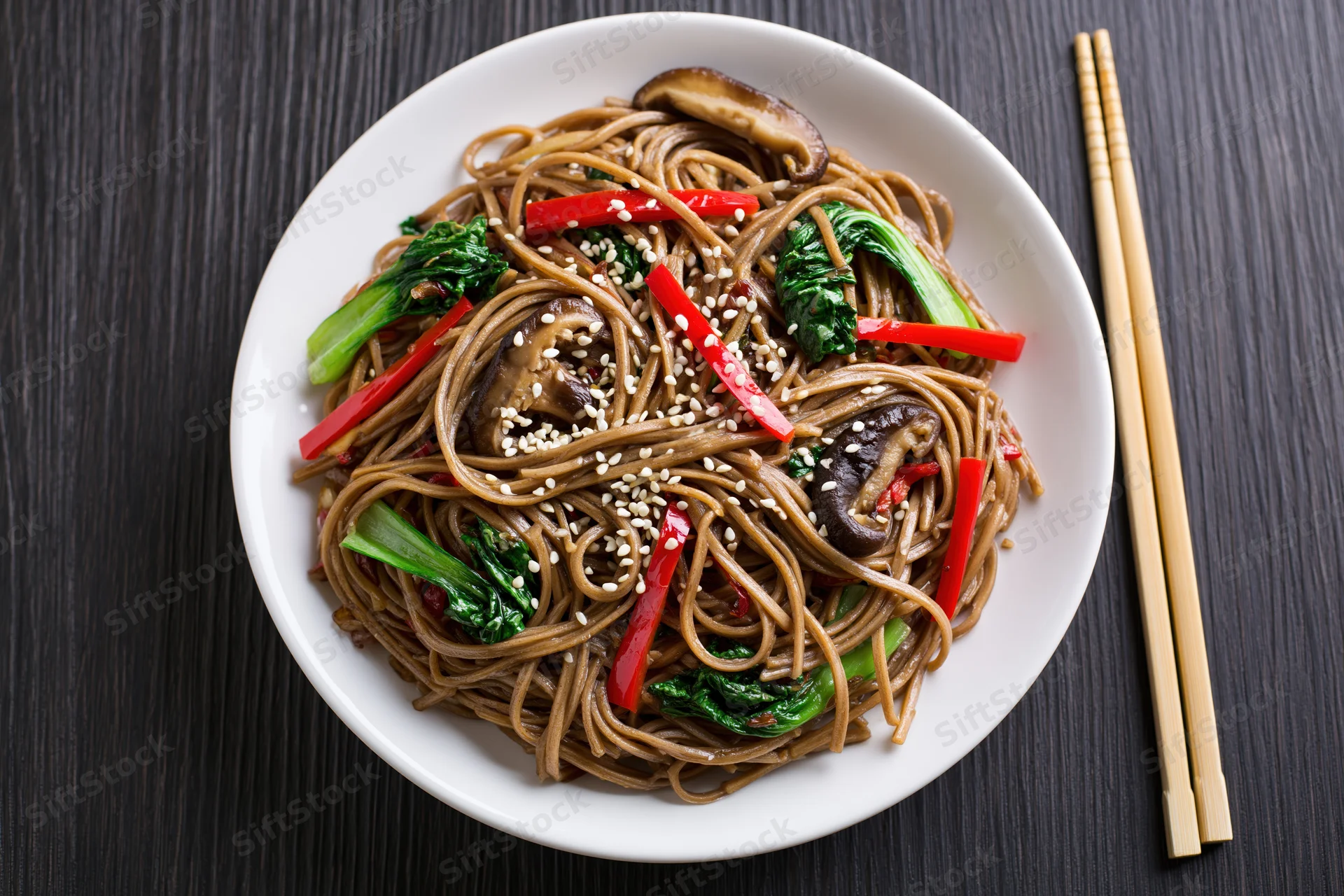 Stir-fried soba noodles with bok choy and shiitake mushrooms on a white plate with chopsticks on a dark wooden table