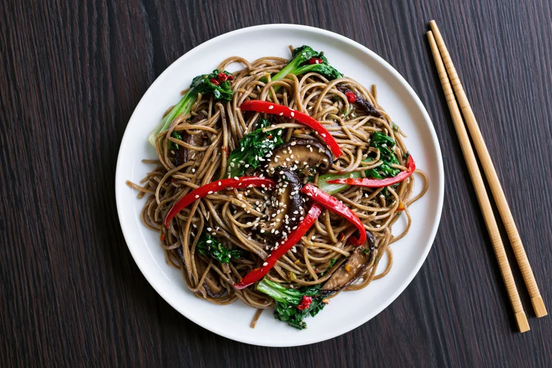 Stir-fried soba noodles with bok choy and shiitake mushrooms on a white plate with chopsticks on a dark wooden table