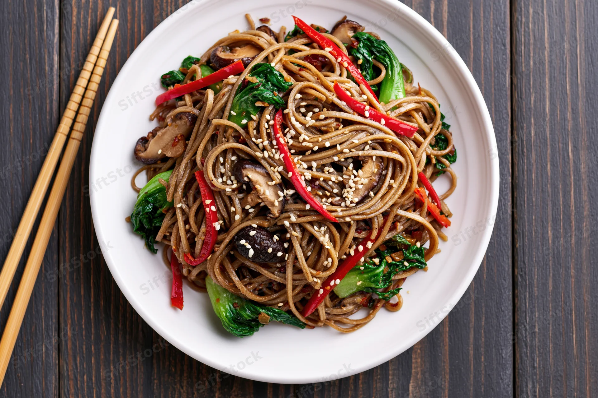 Stir-fried soba noodles with bok choy and shiitake mushrooms on a white plate with chopsticks on a dark wooden table