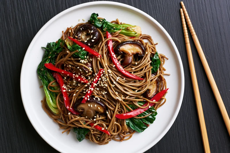 Stir-fried soba noodles with bok choy and shiitake mushrooms on a white plate with chopsticks on dark table