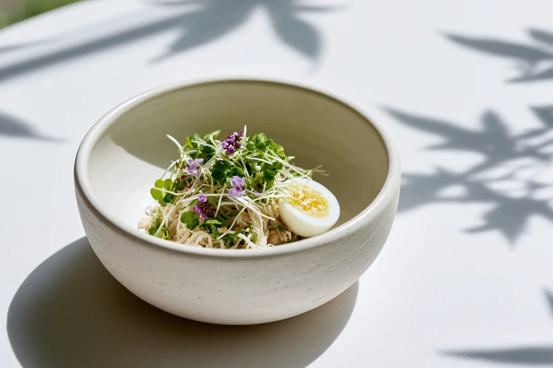 White ceramic bowl with noodles, microgreens, edible flowers and half a hardboiled egg on a sunlit table with leaf shadows