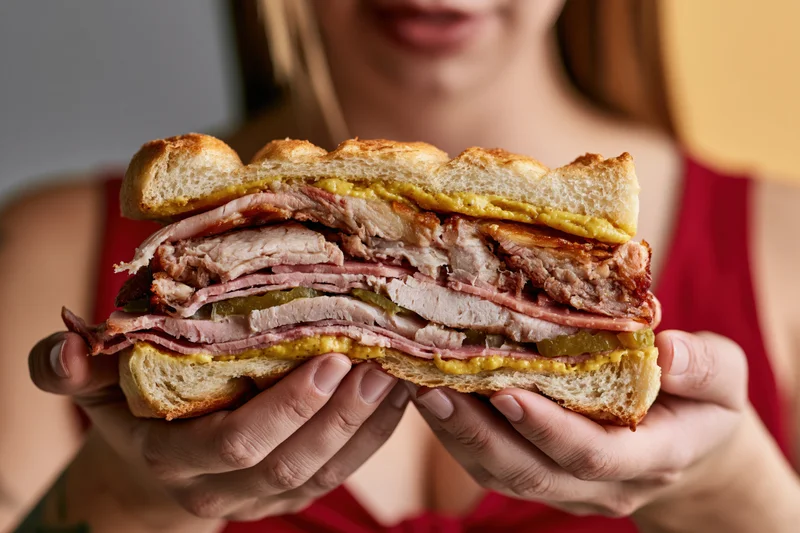 Woman holding a thick cuban sandwich with both hands, layered with ham, pork, cheese, mustard and pickles