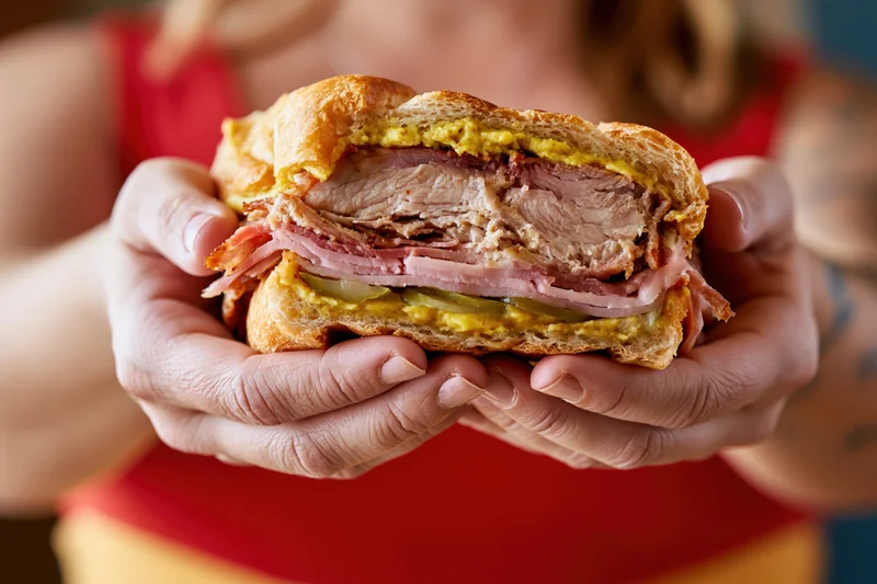 Woman holding a thick cuban sandwich with both hands, showing layers of pork, ham, cheese and pickles