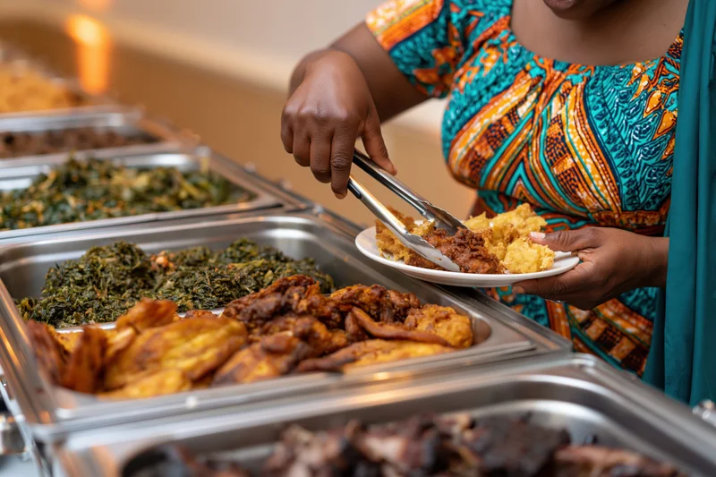 Woman in colorful african print dress serving food from buffet line with tongs and plate
