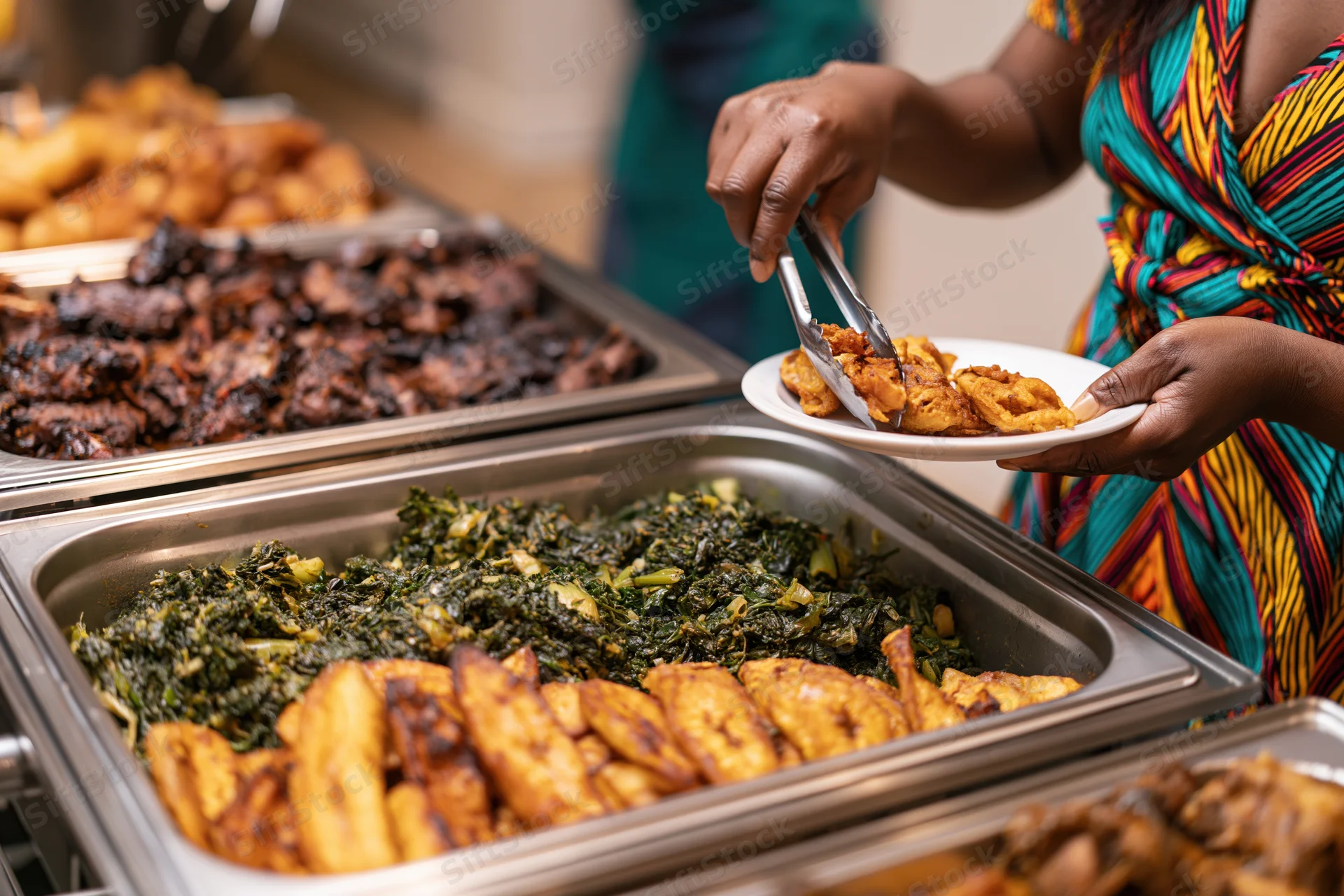 Woman in colorful african print dress serving food from buffet trays with tongs into a plate