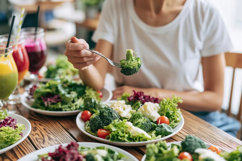 Woman eating fresh garden salad with broccoli and vegetables at wooden dining table
