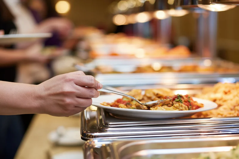 Woman's hand serving food from a hotel buffet line with stainless steel chafing dishes and plates in a warm-lit dining area