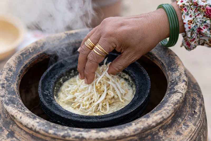 Woman’s hand placing fresh bean sprouts into a steaming clay pot for cooking