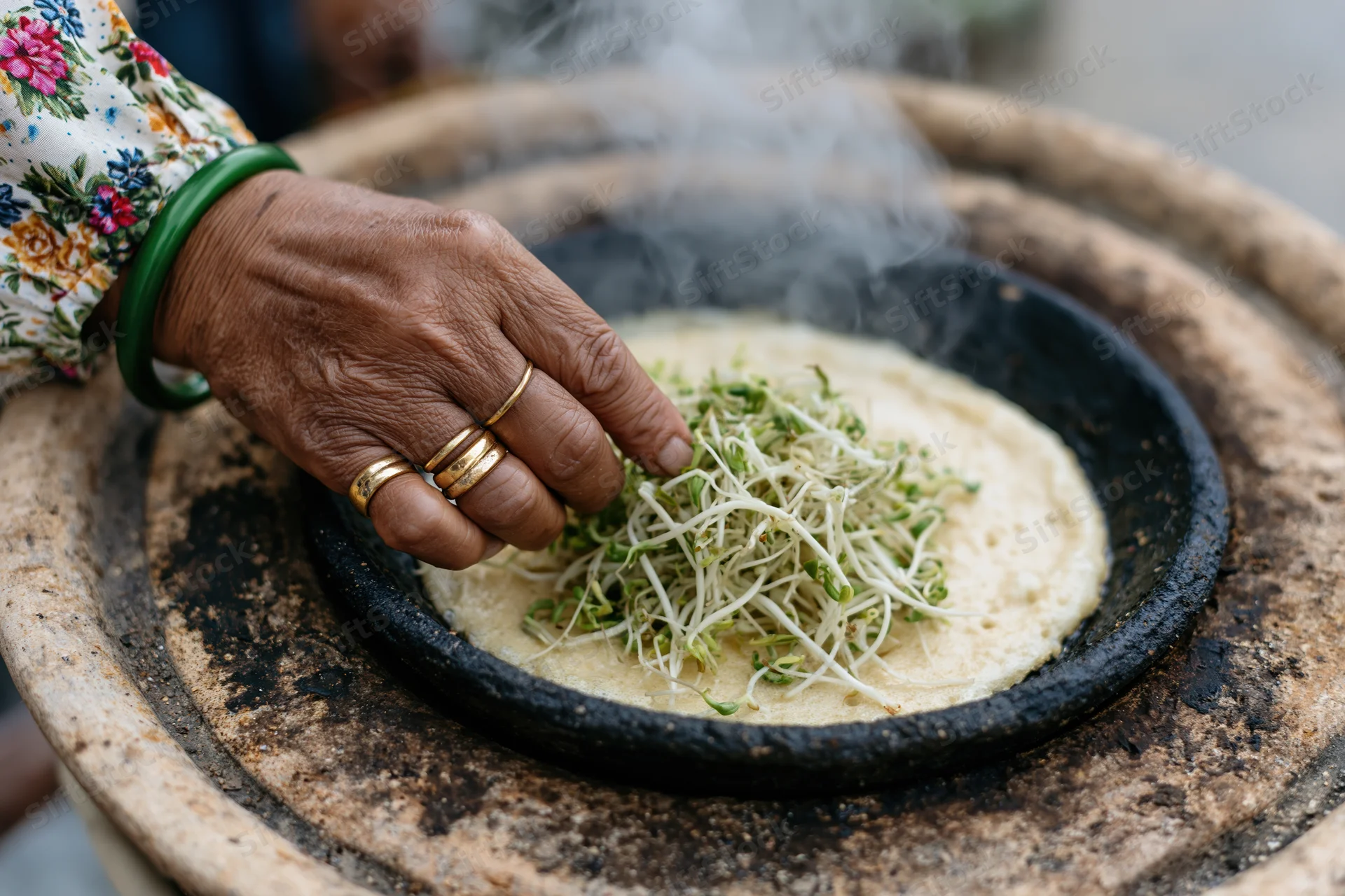 Woman’s hand placing sprouts on steaming flatbread on clay stove