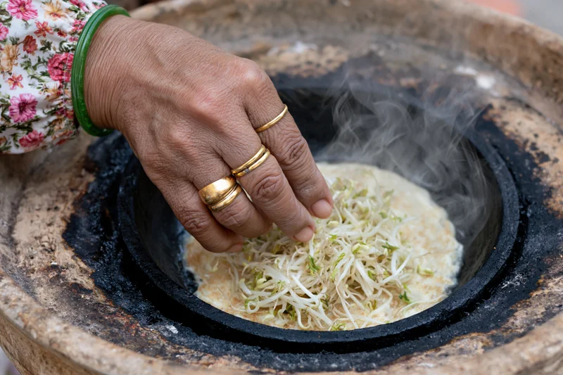 Woman’s hand placing bean sprouts on steaming flatbread in a clay stove