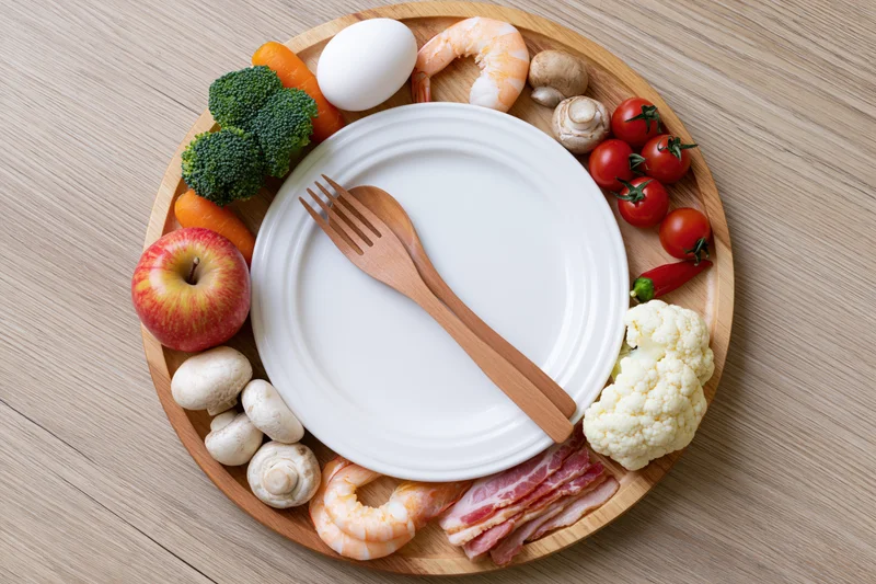 Circular wooden tray with white plate and fork surrounded by various foods arranged like a clock face on a wooden table