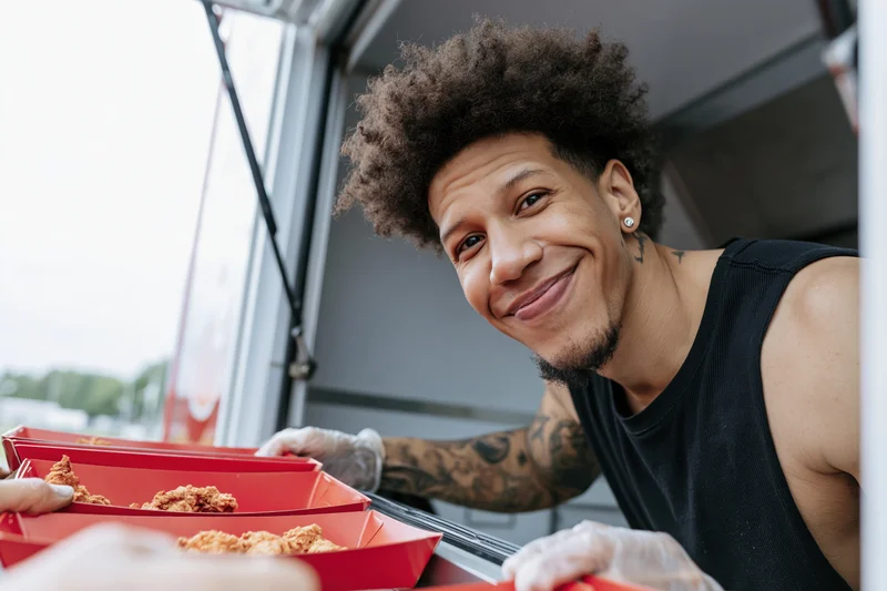 Young black man with afro and tattoos smiling while serving fried chicken from a food truck window