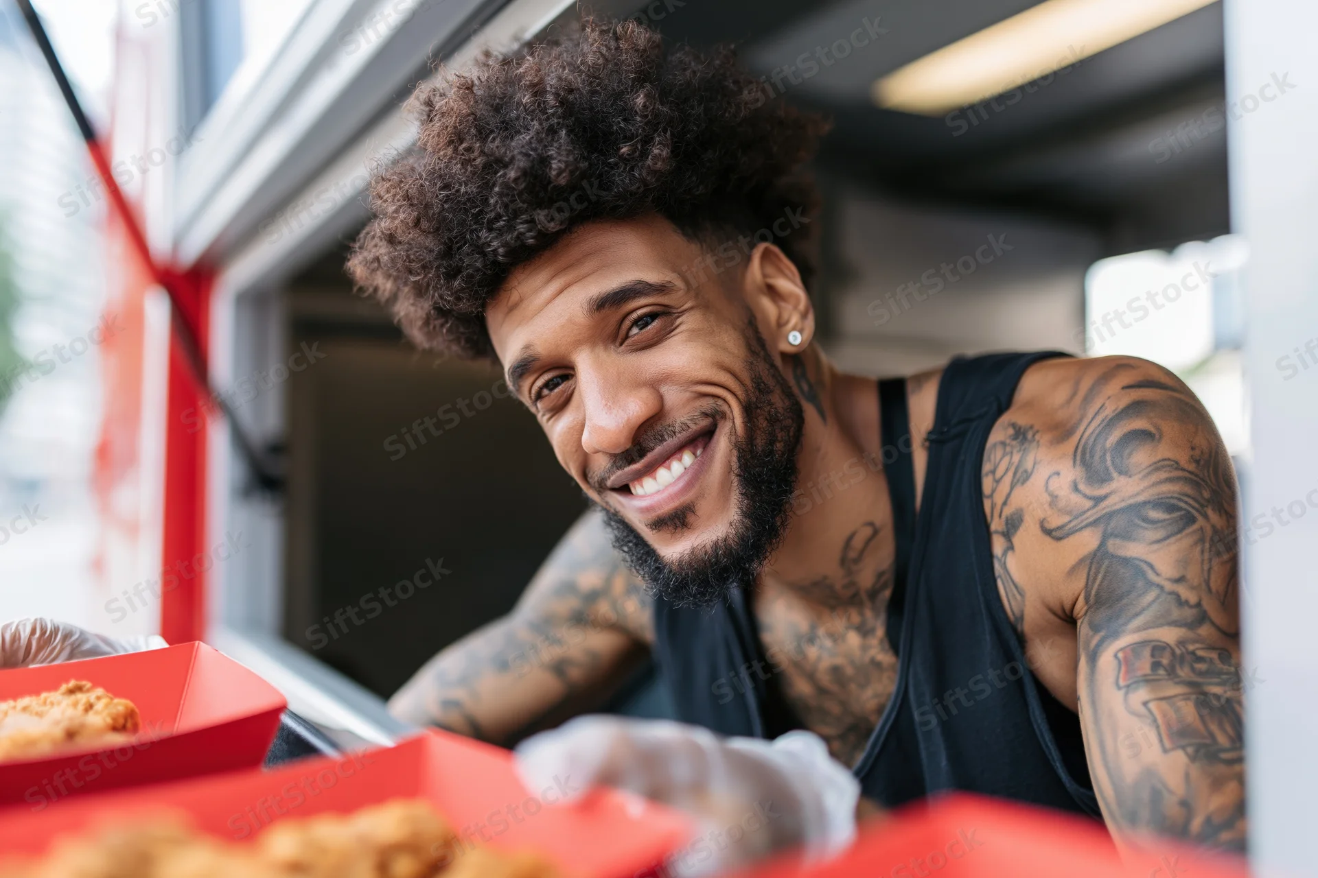 Young black man with afro and tattoos smiling while serving food from a restaurant window