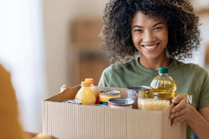 Young black woman smiling while holding a box of donated food items indoors
