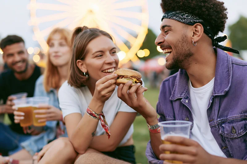 Young woman feeding burger to laughing man at amusement park evening gathering