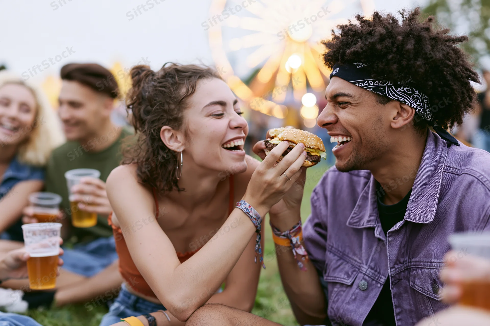 Young woman feeding a burger to a laughing man at an amusement park festival