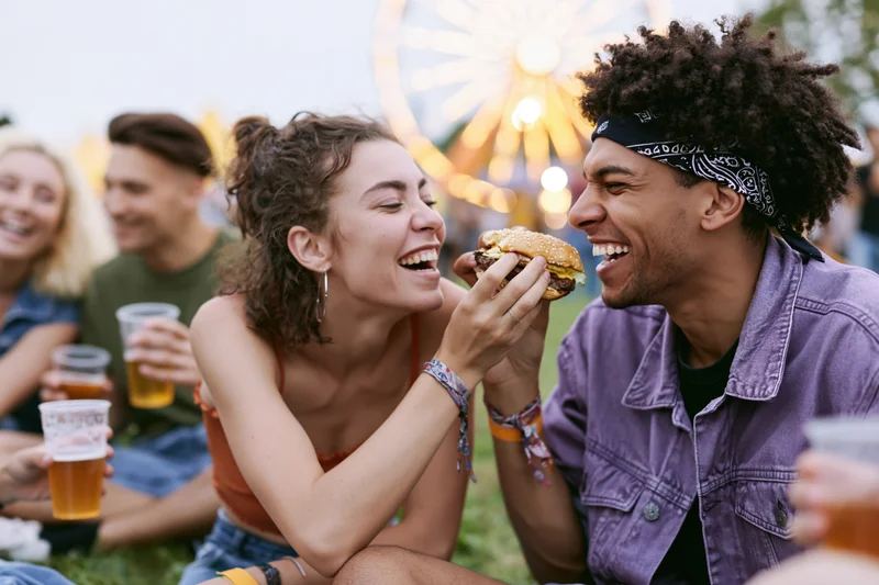 Young woman feeding a burger to a laughing man at an amusement park festival
