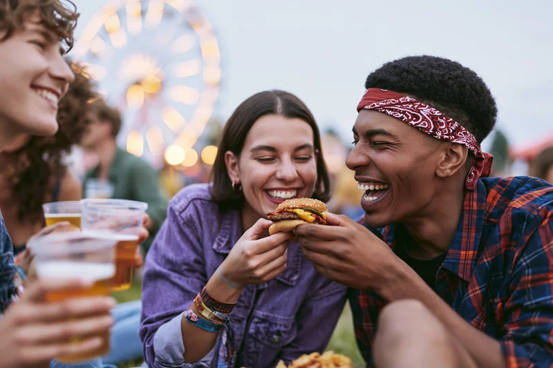 Young woman feeding a burger to a laughing man at an amusement park with ferris wheel in background