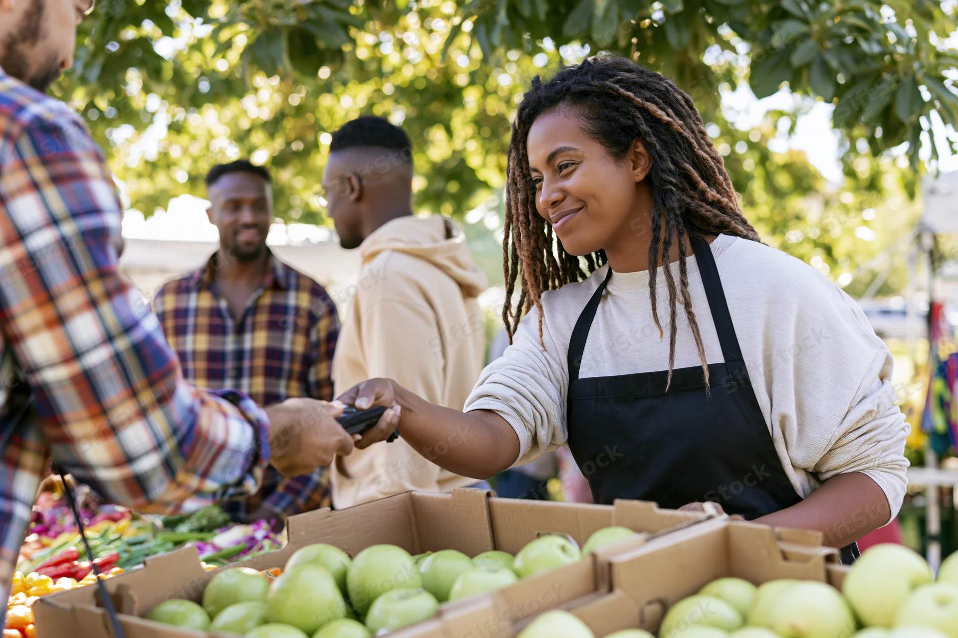 Woman vendor in black apron handing card reader to customer at outdoor produce market