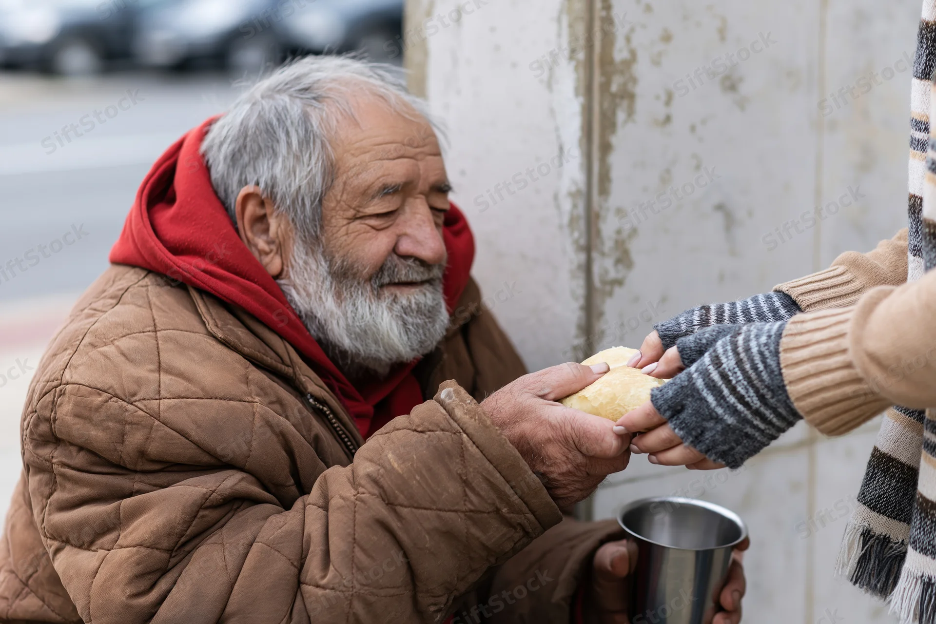 Elderly homeless man receiving bread and metal cup from a helper on a city sidewalk