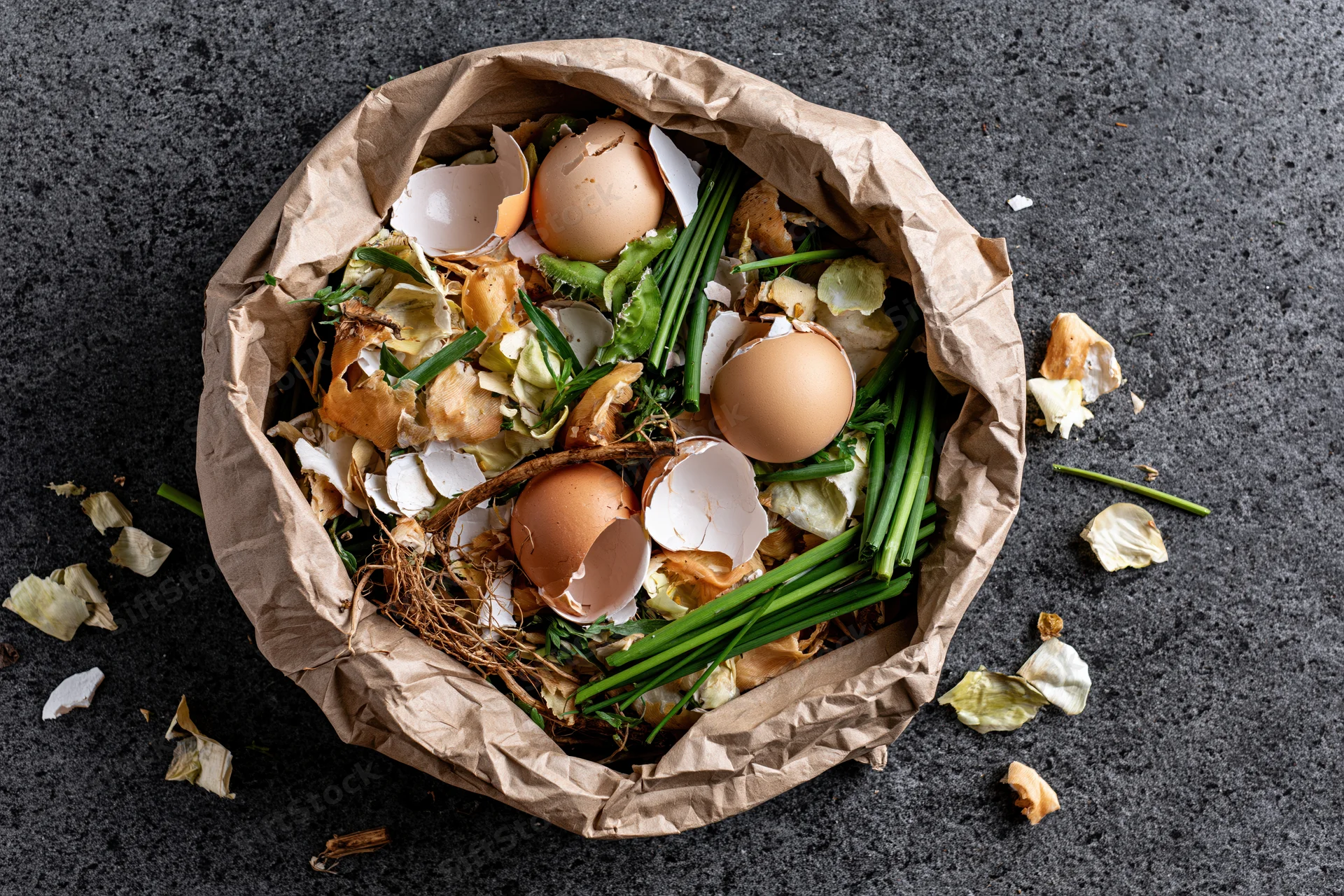 Crumpled brown paper bag filled with kitchen food scraps including eggshells and vegetable peels on dark surface