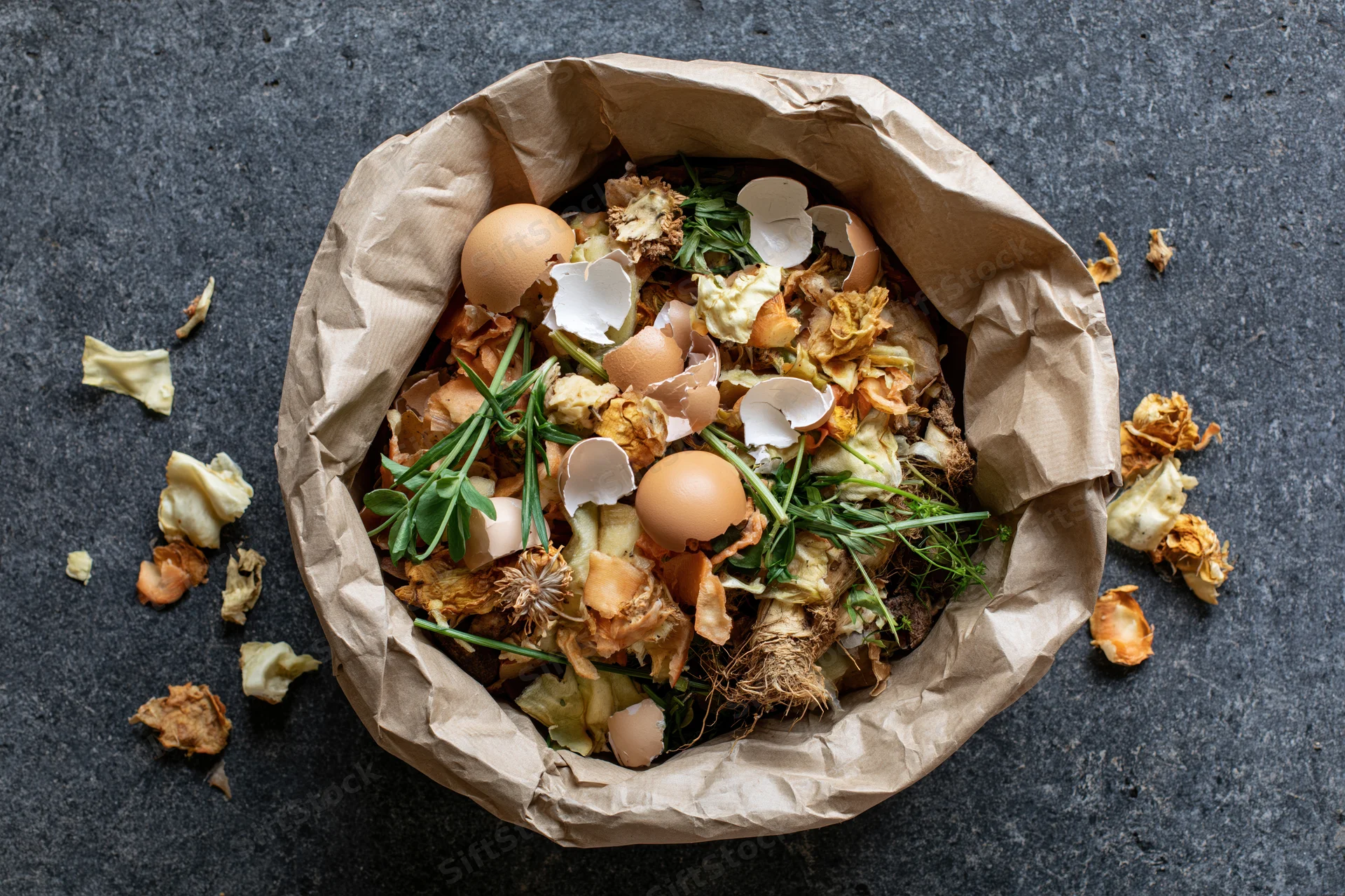 Crumpled brown paper bag filled with kitchen food scraps including eggshells and vegetable peels on dark surface