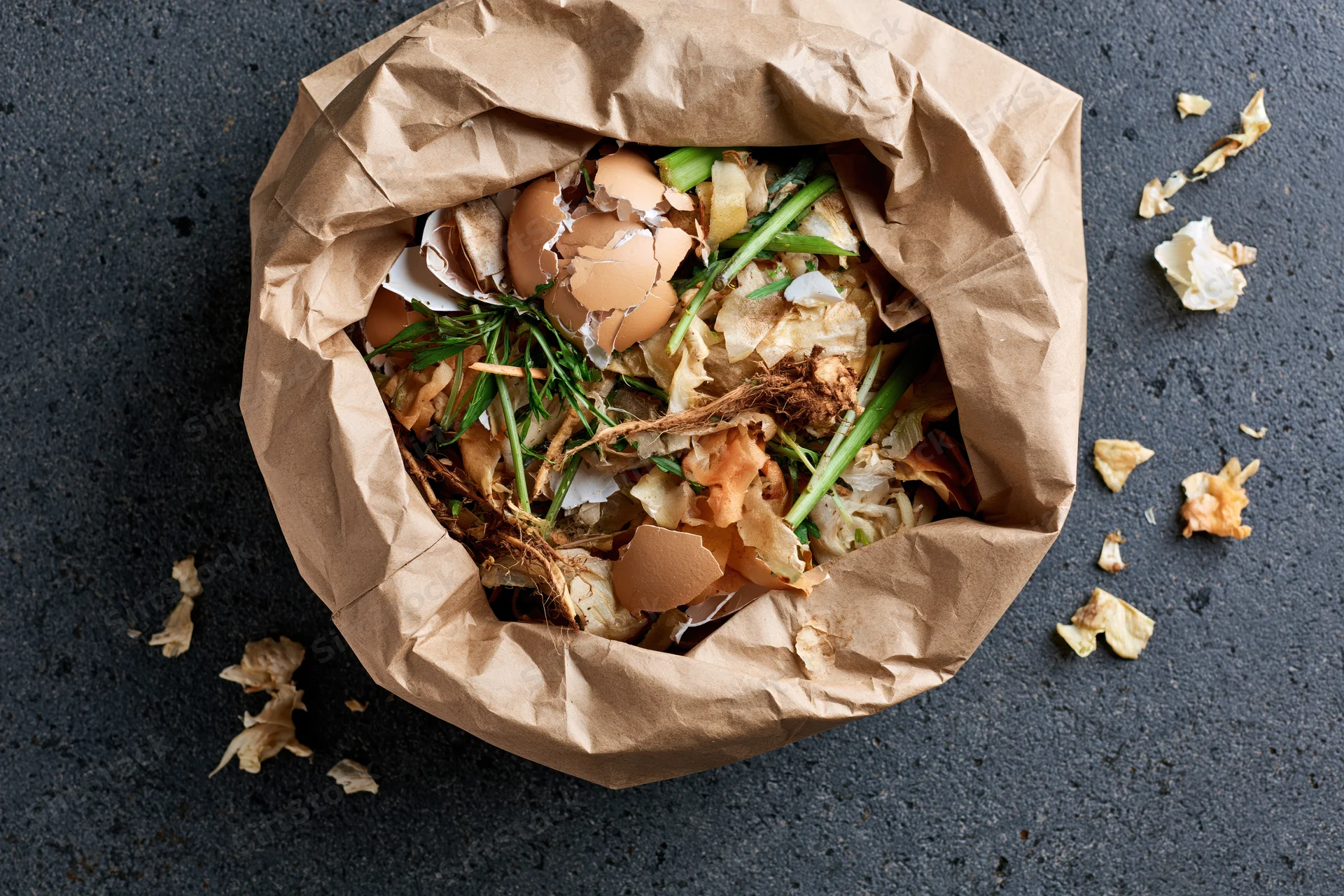 Open crumpled brown paper bag filled with kitchen food scraps on dark surface