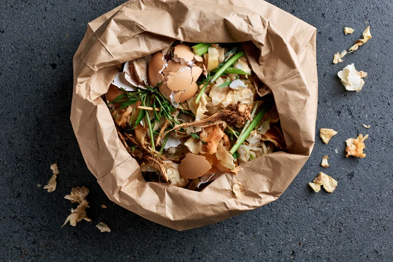 Open crumpled brown paper bag filled with kitchen food scraps on dark surface