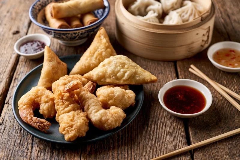 Assorted asian appetizers including dumplings, spring rolls, samosas and tempura shrimp arranged on a rustic wooden table with dipping sauces and chopsticks