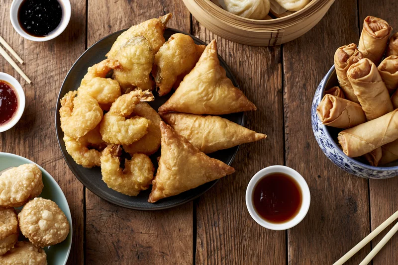 Assorted asian appetizers including samosas, spring rolls, tempura, and dumplings arranged on a rustic wooden table with dipping sauces and chopsticks