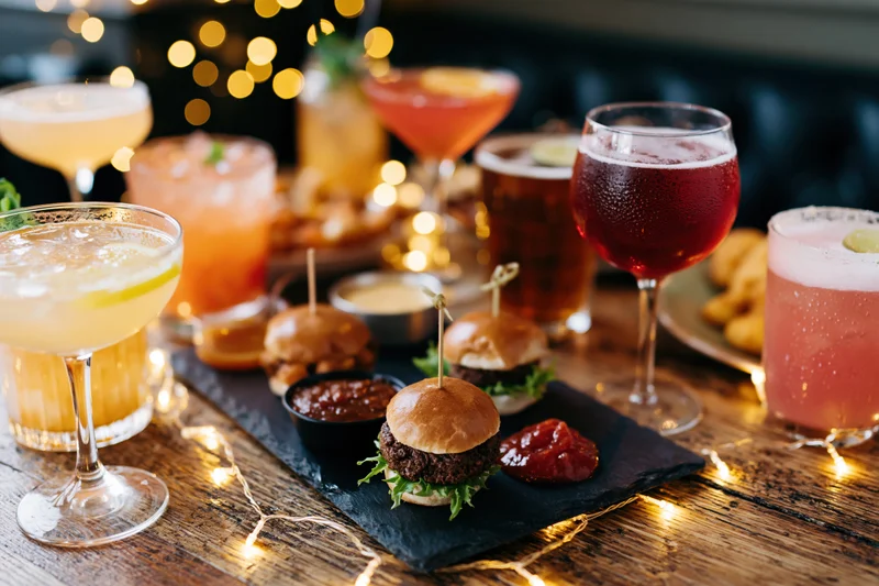 Assorted cocktails and mini burgers with condiments on a rustic wooden table with string lights