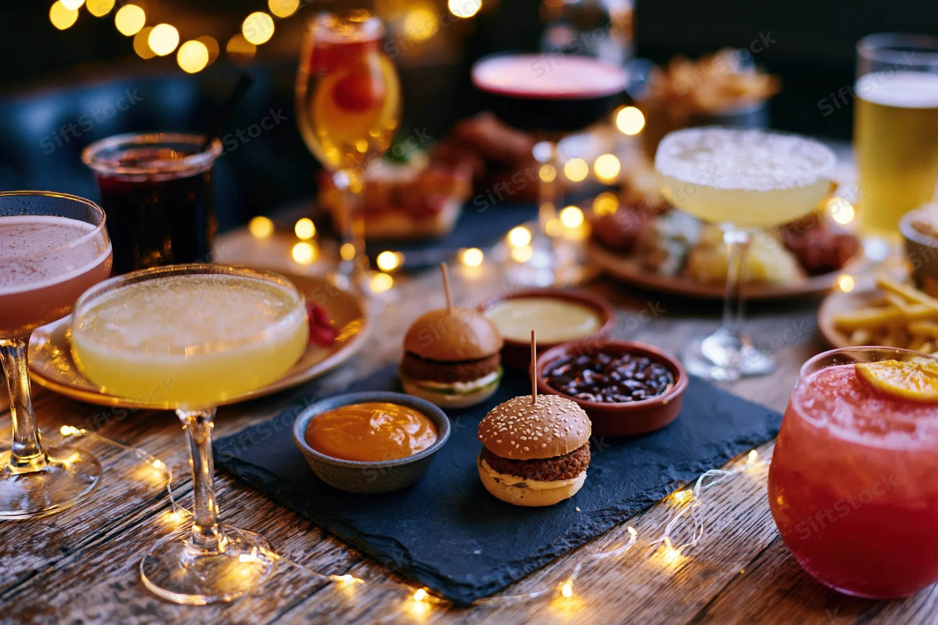 Assorted cocktails and bar snacks arranged on a rustic wooden table with string lights