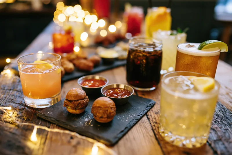 Assorted cocktails and bar snacks arranged on a rustic wooden table with string lights in the background