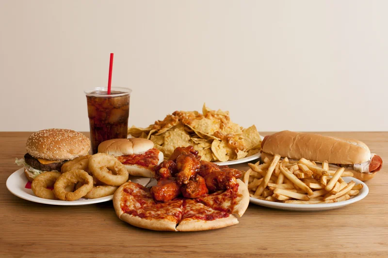 Assorted fast food items arranged on a light wood table with plates and a soda cup