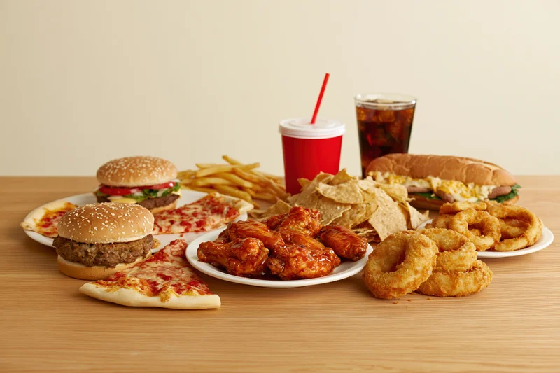 Assorted fast food items including burgers, fries, pizza, chicken wings, onion rings, chips, and soda arranged on a light wood table