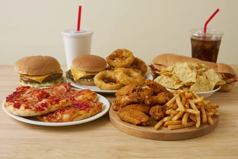 Assorted fast food items including burgers, pizza, fries, chicken wings, onion rings, chips, and soda arranged on a light wood table