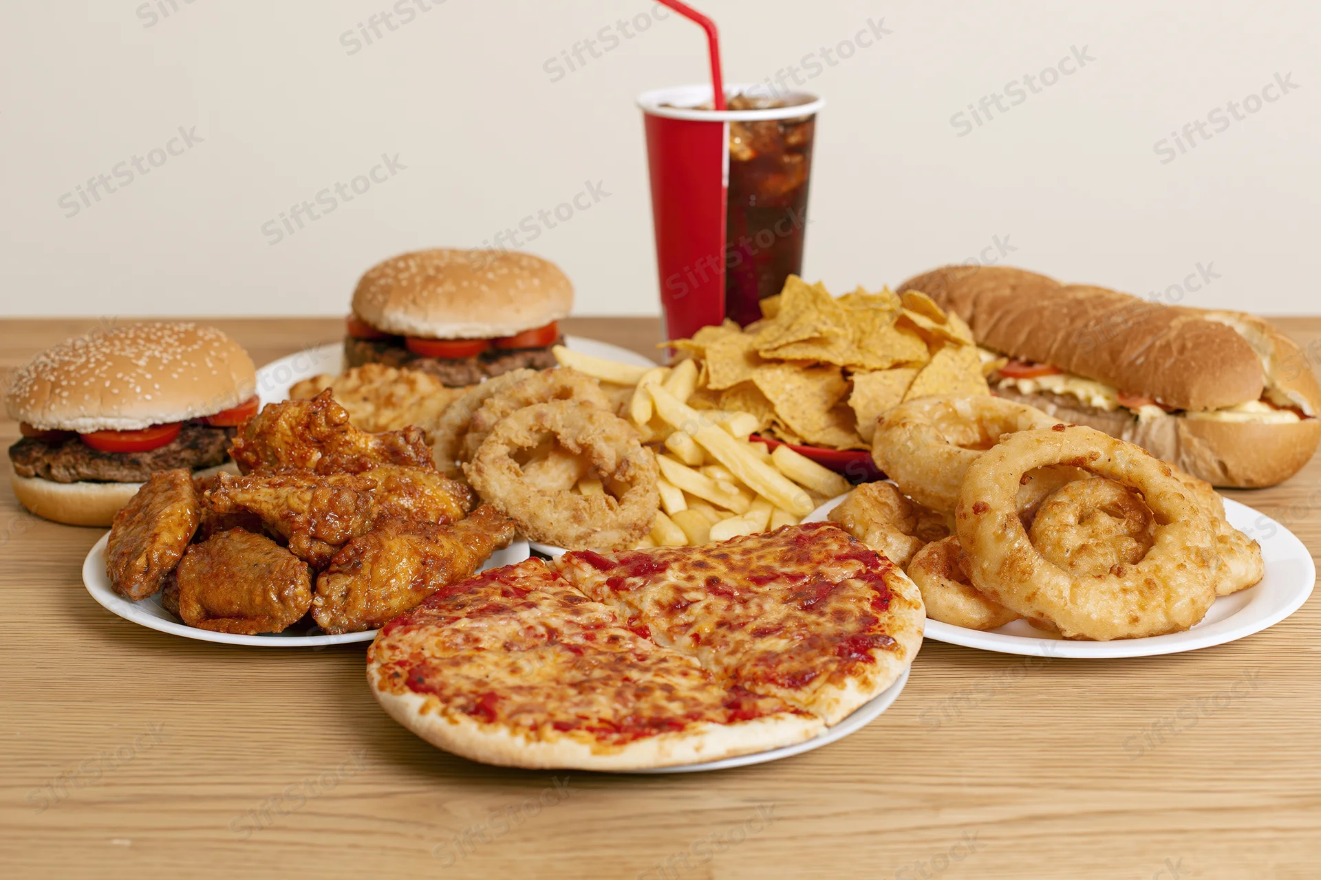 Assorted fast food items arranged on a wooden table including burgers, pizza, fries, onion rings, chicken wings, soda and chips